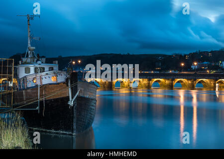 Ein Hausboot sitzt am Ufer des River Torridge in Bideford, wie das Licht auf der langen Brücke in der Abenddämmerung kommen. Stockfoto