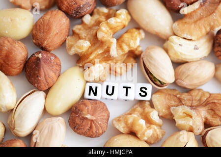 Rohe Nüsse und das Wort "Nuts" von Fliesen- buchstaben Perlen verteilt auf einem weißen Tisch geschrieben Stockfoto