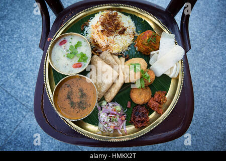 Nahaufnahme der Parsi Hochzeit Thali mit verschiedenen Gerichten serviert auf einer Runde Messing Teller mit Banane Blatt Schonschicht platziert auf einem Holzstuhl gefärbt Stockfoto