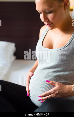 Schwangere Frau sitzt am Bett mit den Händen am Bauch Stockfoto