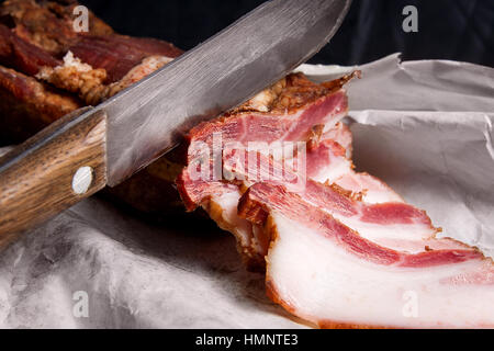 Blick Scheiben geräucherten Speck und Messer auf den weißen Verpackungspapier hautnah. Naturmort im rustikalen Stil auf braune Vintage-Hintergrund. Stockfoto