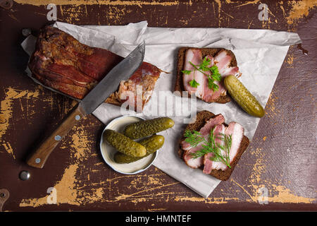 Nahaufnahme Blick Scheiben geräucherter Speck auf das Stück Roggenbrot mit Kräutern, mehrere Gurken, altes Messer mit Holzgriff - rustikalen Stil auf braun vintage Stockfoto