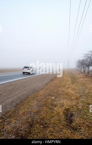 Eine silberne Fahrzeug fährt auf eine asphaltierte Landstraße in einem nebligen Morgen in Südwest-Ontario, Kanada. Stockfoto