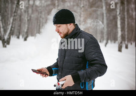 ein junger Mann mit einem Bart, raucht eine elektronische Zigarette und glücklich das Telefonieren über das Internet, eine positive entspannen im Freien in der sn Stockfoto