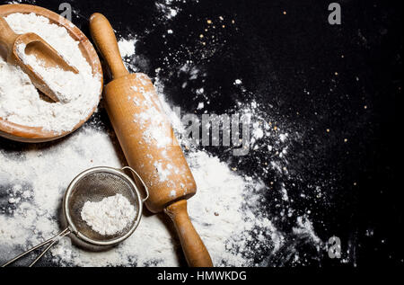 Metall Sieb, Nudelholz und hölzerne Schüssel mit Mehl auf einem schwarzen Hintergrund zu verschütten. Stockfoto