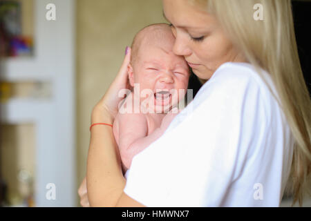 Junge Mutter wiegt und beruhigende Lied singt, sanft umarmen, küssen und im Gespräch mit Baby, Neugeborene runzelt die Stirn und Weinen befindet sich an Mama Händen im Licht ro Stockfoto