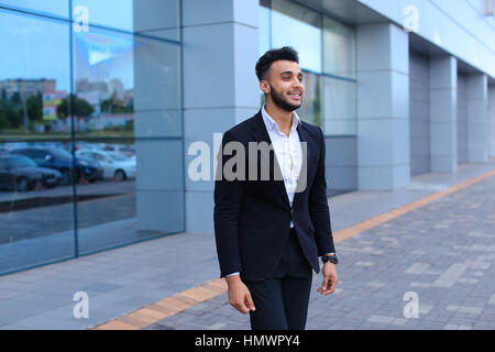 Junge attraktive arabischen männlichen Mann lächelnd, steht und freut sich auf Grund des grauen Business-Center mit großen Fenstern. Muskulöser bärtigen Mann gekleidet Stockfoto