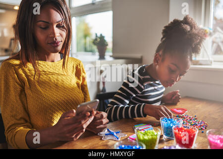 Beschäftigt junge Frau in gelb überprüft ihr Telefon während Kind Perlen aus verschiedenen Farben auf Tisch wählt Stockfoto