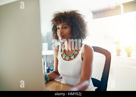 Ziemlich stylish junge afroamerikanische Frau arbeitet an einem großen Desktop-Monitor in einem hohen zentralen Büro lesen am Bildschirm Stockfoto