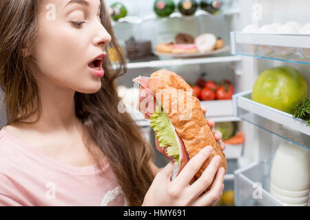 Junge Frau, die große Sandwich vor der Kühlschrank voller Kalorienniveau und Gemüse zu essen Stockfoto