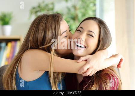 Liebevolle Mädchen küssen ihre glückliche Schwester oder Freundin im Wohnzimmer zu Hause mit einem heimeligen Hintergrund Stockfoto