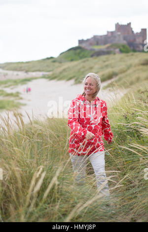 Mittleren Alter Frau zu Fuß auf der Düne mit einer Burg im Hintergrund. Sie ist warme Bekleidung und Lächeln auf den Lippen tragen. Stockfoto