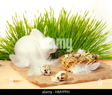 Wachteleier mit Stroh und Federn in Korb, weiße Hase auf Sackleinen und Frühlingsgrün Rasen, Ostern-Konzept Stockfoto