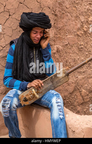 Marokko.  Teenager Amazigh Berber Musiker mit ein Gimbrie, auf seinem Handy zu sprechen.  Ait Benhaddou Ksar, ein UNESCO-Welterbe. Stockfoto