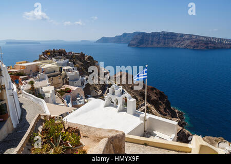 Griechische Flagge durch den Glockenturm der weiß getünchten griechischen orthodoxen Kirche mit Blick auf Thirassia in Oia, Santorini, einer griechischen Mittelmeerinsel Stockfoto