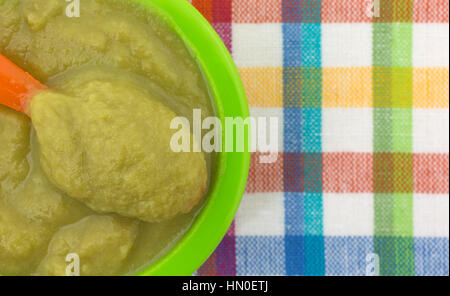 Enge Draufsicht einer Portion Erbsen Babynahrung in einer kleinen Schüssel mit einem Plastiklöffel orange auf einem bunten Tischset. Stockfoto