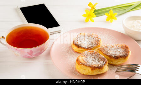 Käse-Pfannkuchen mit Puderzucker, schwarzer Tee, Blumen und Smartphone auf einem weißen Holztisch. Stockfoto