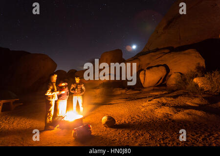 Drei Freunde stehen für Wärme an einem kalten und windigen Nacht im Joshua Tree National Park in der Nähe des Lagerfeuers. Stockfoto