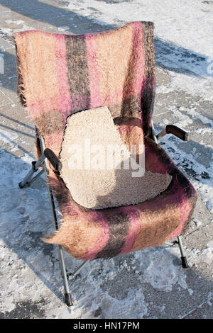 Alte, gebrauchte Aluminium-Metall-Stuhl unter freiem Himmel auf dem Schnee bedeckt Steinpflaster. Der Stuhl ist mit einem Wolltuch bedeckt. Kaltes Wetter Stockfoto