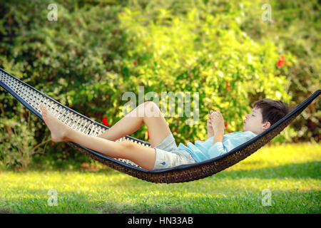 Seitenansicht des niedlichen kleinen Jungen liegen auf einer Hängematte mit Blick auf sein Handy und entspannend Stockfoto