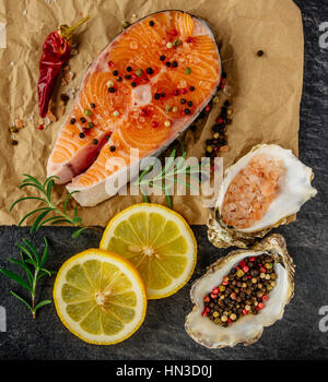 Leckeres Lachssteak, reich an Omega-3 Öl, mit aromatischen Kräutern und Gewürzen mit Zitrone, Tomate, Knoblauch auf schwarzem Hintergrund. Ernährung und gesund essen. Stockfoto