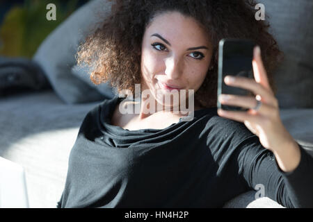 Natürliche entspannt attraktive junge Frau unter ihr Selfie auf einem Smartphone, wie sie zu Hause auf der Couch entspannt sich Stockfoto