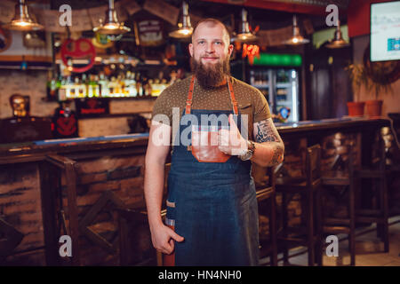 Bärtige Barkeeper mit Tattoos und Uhren tragen eine Schürze Stand in der Nähe der Bar. Stockfoto