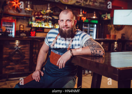 Bärtige Barkeeper mit Tattoos und Wathes trägt eine Schürze sitzt in Bar. Stockfoto