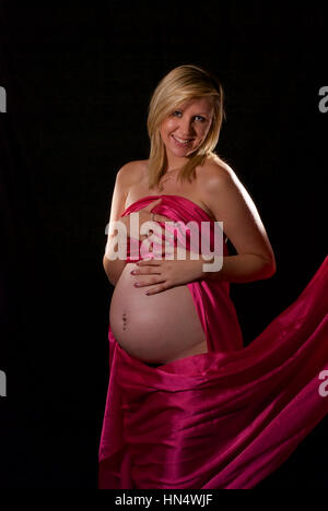 Schöne blonde behaarte schwangere junge Frau in Rosa Seide wickeln Stockfoto