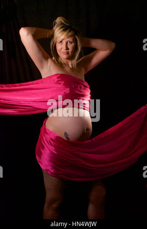 Schöne blonde behaarte schwangere junge Frau in Rosa Seide wickeln Stockfoto