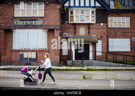 Junge Mutter schieben Kinderwagen letzten außen geschlossen und mit Brettern vernagelt Windows Grün Ende eine großen Wahrzeichen Kneipe in Burgage, South Manchester, England, UK, Stockfoto