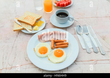 Frühstück mit gebratenes Ei, Schinken, Wurst, Toast, Marmelade, Butter, Wassermelone, Orangensaft und schwarzen Kaffee auf Marmortisch. Stockfoto