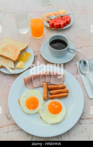 Frühstück mit gebratenes Ei, Schinken, Wurst, Toast, Marmelade, Butter, Wassermelone, Orangensaft und schwarzen Kaffee auf Marmortisch. Stockfoto