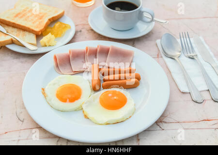 Frühstück mit gebratenes Ei, Schinken, Wurst, Toast, Marmelade, Butter, Wassermelone, Orangensaft und schwarzen Kaffee auf Marmortisch. Stockfoto