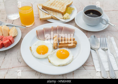 Frühstück mit gebratenes Ei, Schinken, Wurst, Toast, Marmelade, Butter, Wassermelone, Orangensaft und schwarzen Kaffee auf Marmortisch. Stockfoto