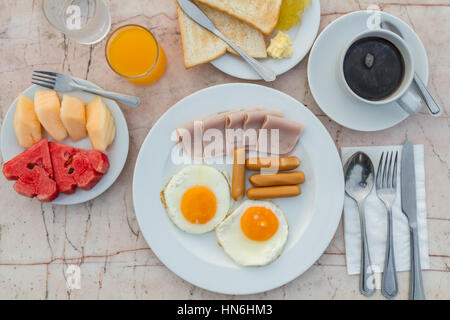 Frühstück mit gebratenes Ei, Schinken, Wurst, Toast, Marmelade, Butter, Wassermelone, Orangensaft und schwarzen Kaffee auf Marmortisch. Ansicht von oben. Stockfoto