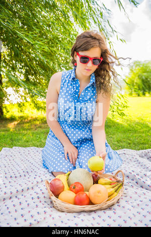 Junges Mädchen im Kleid und rote Sonnenbrille sitzt auf Picknick Decke Kommissionierung grüner Apfel vom Obstkorb Stockfoto