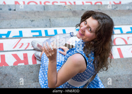 Junge Frau mit blau gepunkteten Kleid sitzt auf Schritte in eine klare Flasche stilles Wasser trinken und lachen Stockfoto