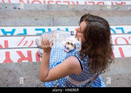 Junge Frau mit blau gepunkteten Kleid sitzt auf Schritte in eine klare Flasche stilles Wasser trinken und lachen Stockfoto