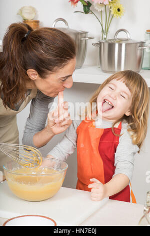 drei Jahre alt Schlagsahne Kind Lachen und Zunge mit Mutter saugen ihre Finger nach Belieben herausnehmen, in Teamarbeit, Kochen ein Biskuit bei kitc Stockfoto