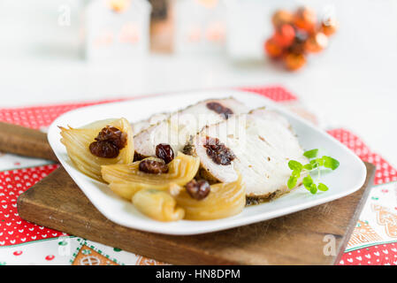 Scheiben vom gebratenen Schweinekoteletts gefüllt mit Preiselbeeren und Knoblauch mit gekochten Zwiebeln und frischer Oregano auf weißen Teller auf rustikale Schneidbrett angeordnet. Stockfoto