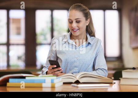 Junger Student Studium in der Bibliothek, sie ist SMS und apps auf ihrem Smartphone verwenden Stockfoto