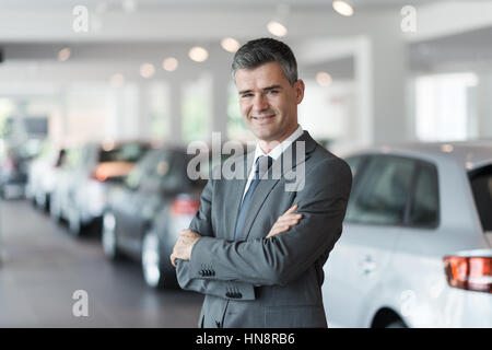 Zuversichtlich Lächeln Autoverkäufer im Showroom, steht er mit verschränkten Armen und Luxus-Autos auf dem Hintergrund Stockfoto