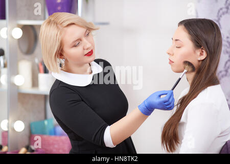 Professionelle Visagistin tragen schwarzes Kleid und Handschuhe machen, attraktive junge Frau mit reiner Haut bilden. Blonde professionelle Verfahren Pinsel Stockfoto