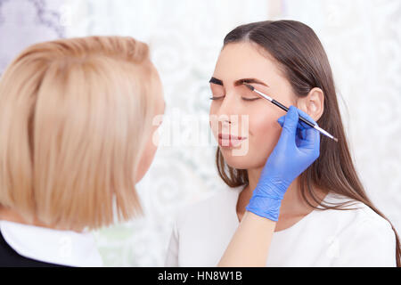 Porträt von professionellen blonde Kosmetikerin in blaue Handschuhe machen permanente Augenbrauen für Frau, mit Pinsel. Weiblichen Kunden mit geschlossenen Augen Lächeln Stockfoto