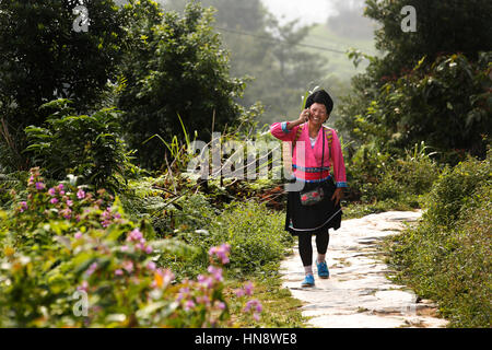 Eine Frau im traditionellen Yao Outfit spricht auf dem Handy wie geht sie entlang eines Pfades durch die Longji Rice-Terrassen in der Nähe von Guilin, Provinz Guangxi, Stockfoto