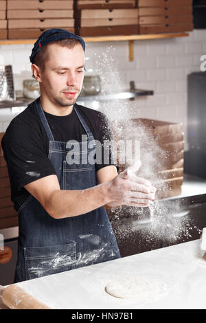 Den Teig ausrollen. Wolke aus Mehl erweitert. Closeup Hand des Chef-Bäcker in einheitliche blaue Schürze Koch Pizza in der Küche Stockfoto