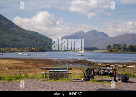 Loch Leven, Blick nach Westen von Glencoe Village mit Backpacker auf der Bank mit Rucksack neben seinem Kopf hinlegen. Stockfoto