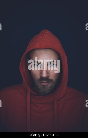 Porträt eines Weißen, Mitte dreißig, im Studio erschossen, mit einem roten Kapuzenpulli an einer dunkelblauen Wand. Stockfoto