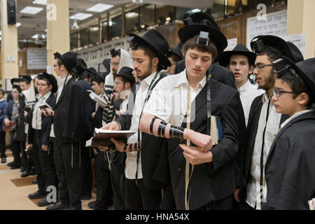 Orthodoxe jüdische Jüngling aufsetzen Gebetsriemen vor morgen Dienstleistungen in einem überfüllten Synagoge in Brooklyn, New York. Stockfoto